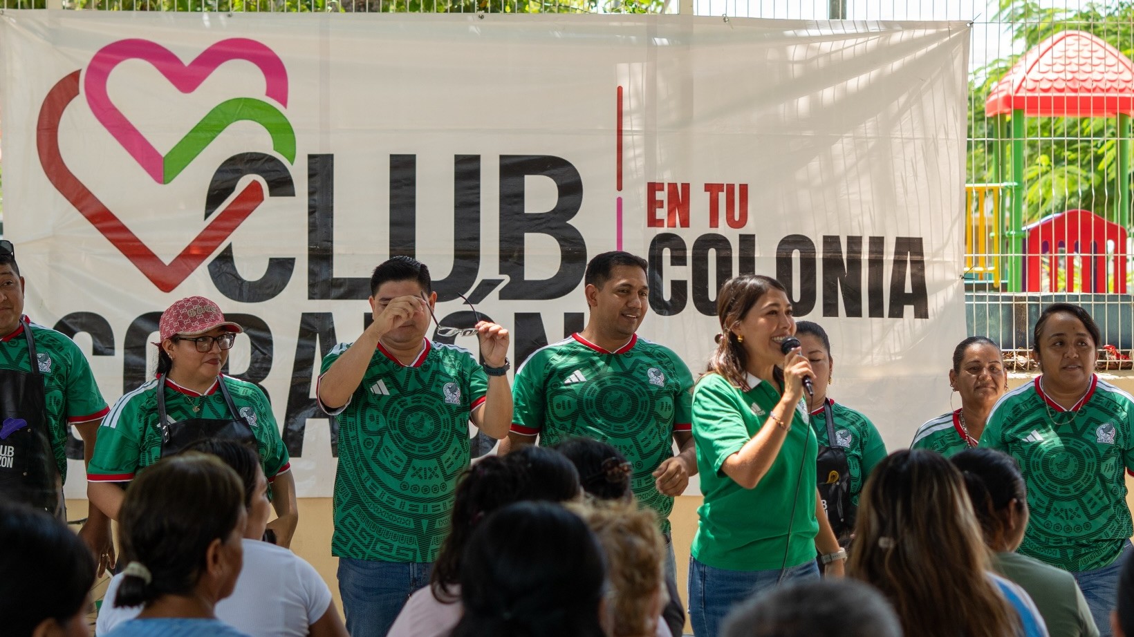 Mirella habla al micrófono frente a la lona de Club Corazón en tu Colonia, con el equipo a su lado y los vecinos sentados escuchándola